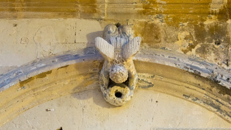 Weathered carved stone creature with an open circular spout projecting above the curved molding of an arch.