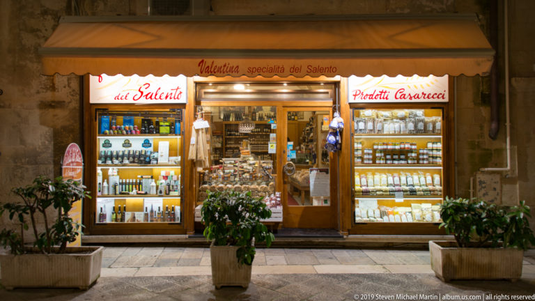 Illuminated Salento specialty-food shop with shelves of bottles and packaged goods behind two display windows and potted plants outside.