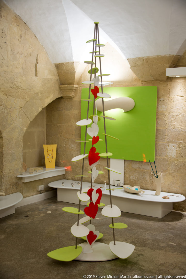 Abstract metal tree-like installation with white discs and red hearts in a vaulted stone room with green display panels.