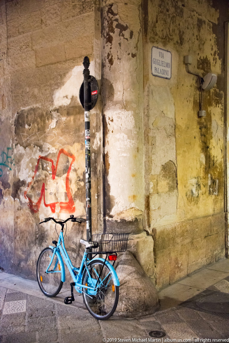 Blue bicycle leaning against a weathered stone corner beneath a Via Guglielmo Paladini street sign and traffic signal.