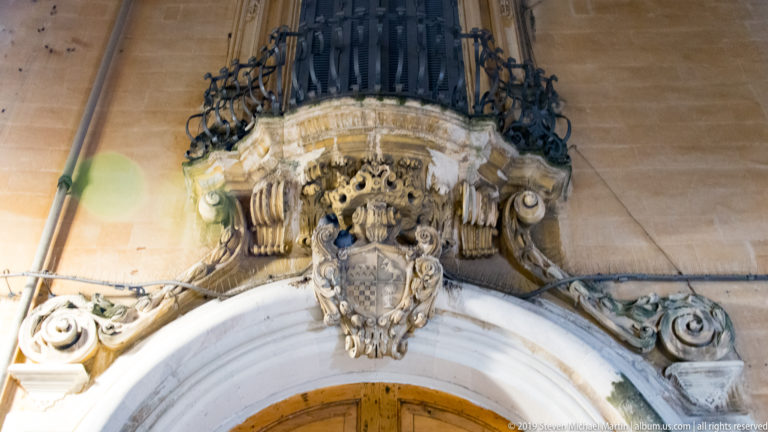 Ornate carved coat of arms above an arched doorway, directly beneath a projecting curved wrought-iron balcony.