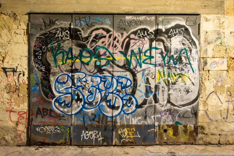 Dark double doors densely covered in colorful graffiti tags, set within a rough pale-stone wall also marked with paint.