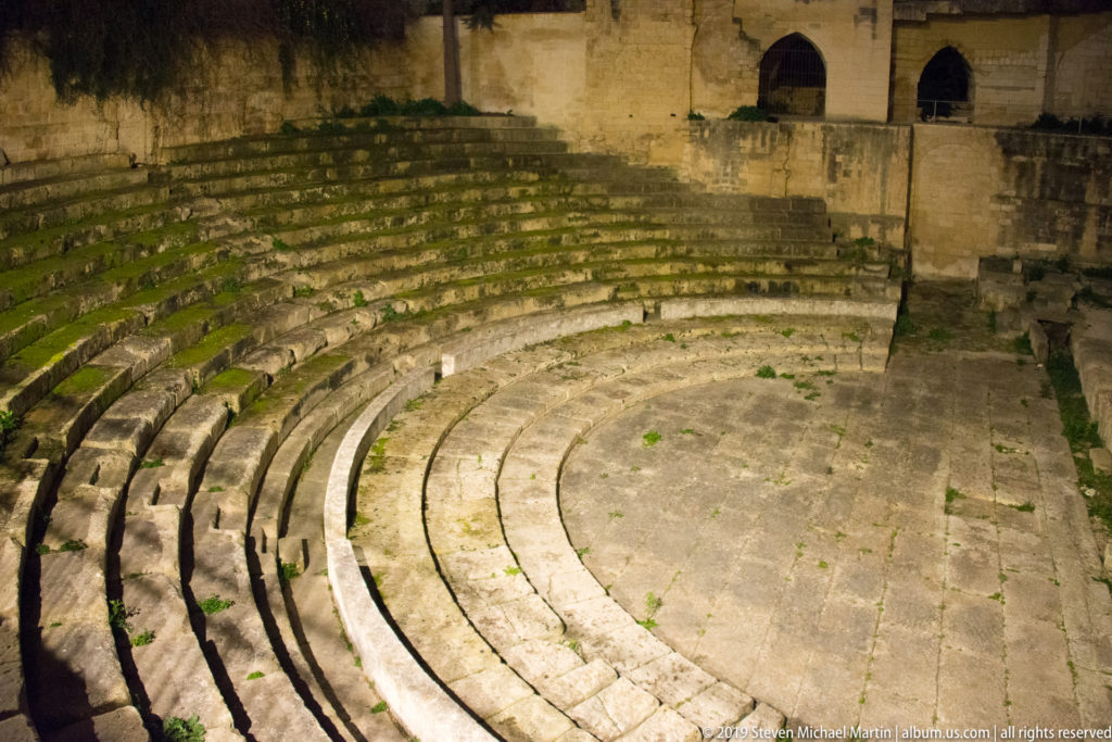 Curving tiers of ancient stone seating descending toward the arena, with arched openings in a high wall beyond.