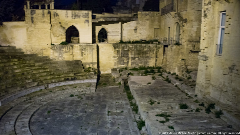 Curved stone arena and stepped seating of Lecce’s Roman amphitheatre with arched openings in the illuminated perimeter wall.