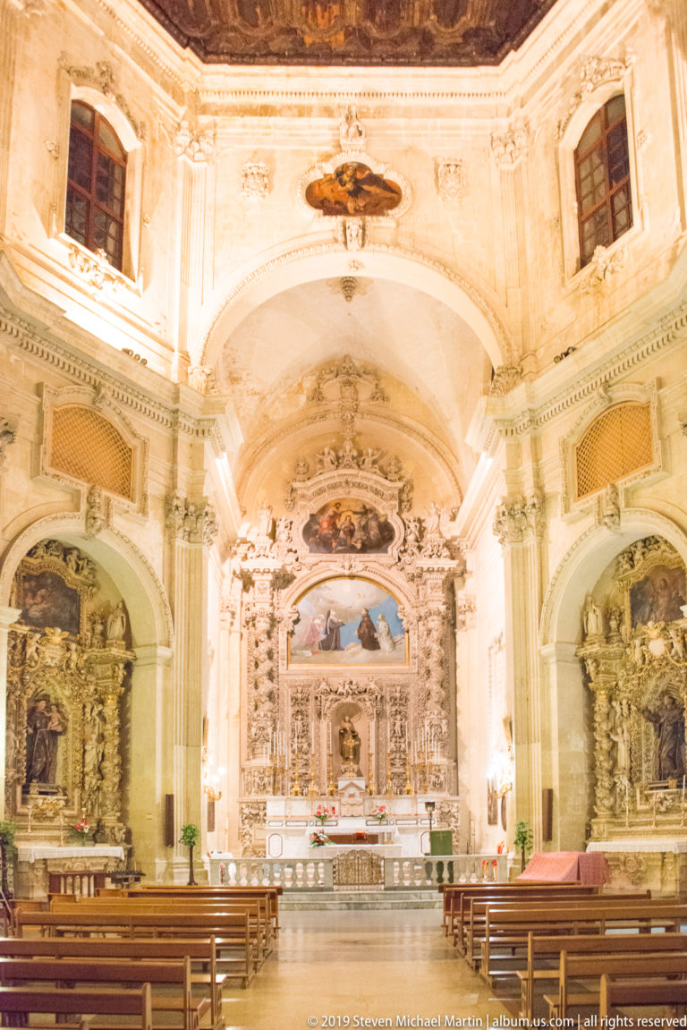 Wide church interior with wooden pews leading toward an ornate high altar beneath a decorated vault and flanked by side chapels.