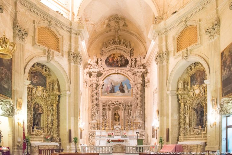 Bright Baroque church nave facing an elaborate high altar, with ornate side chapels, paintings, arches, and a vaulted ceiling.