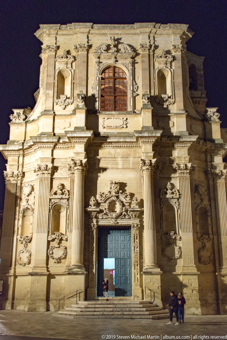 Highly ornate illuminated stone church façade with paired columns, statues in niches, relief carving, central doorway, and upper window.