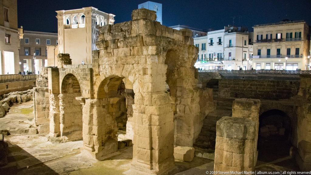 Tall ancient masonry remnant with multiple arches and heavy piers rising from Lecce’s Roman amphitheatre excavation at night.