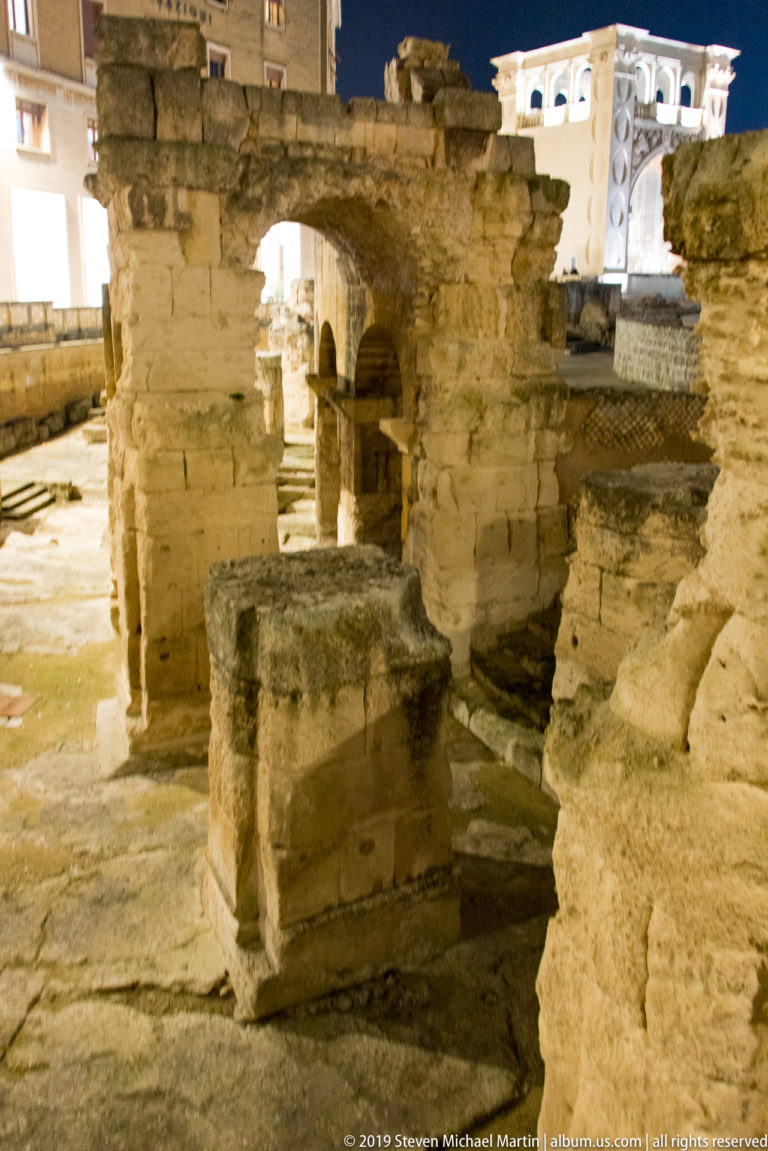 Tall arched masonry remains of Lecce’s Roman amphitheatre rising above the excavation with Piazza Sant’Oronzo illuminated beyond.