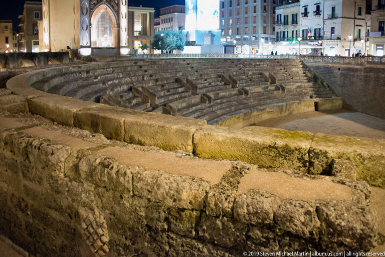 Foreground stone blocks frame an oblique nighttime view of the Roman amphitheatre’s curved seating tiers and surrounding city buildings.
