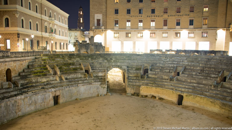 Roman amphitheatre seating and central vaulted passage at Piazza Sant’Oronzo with the Palazzo Carafa inscription visible behind.