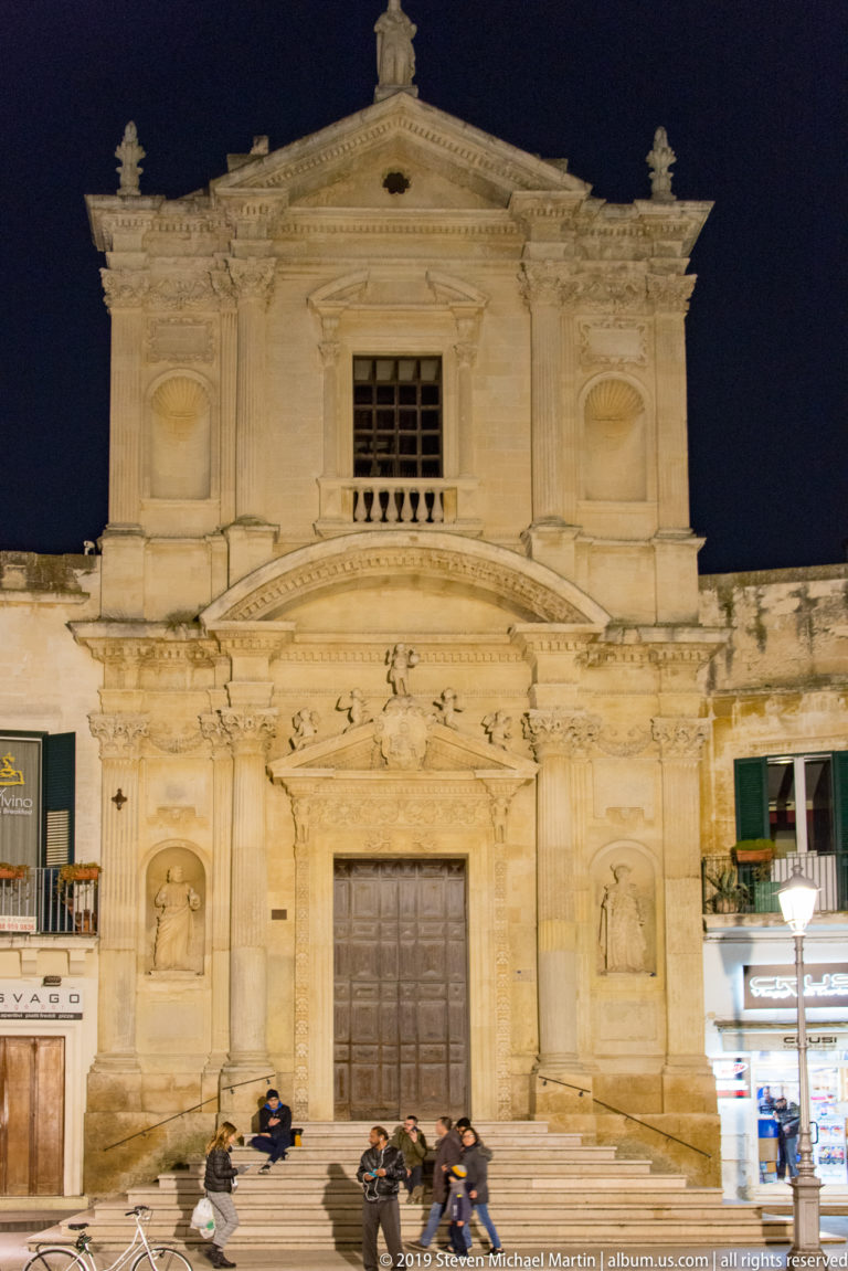 Illuminated pale-stone church façade with central door, niches, pilasters, upper pediment, and people seated on the steps.