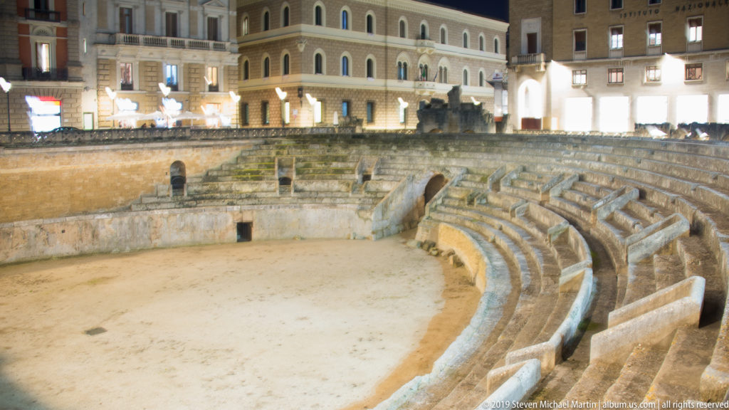 Curved stone seating and arena floor of Lecce’s Roman amphitheatre, surrounded by illuminated modern buildings at night.