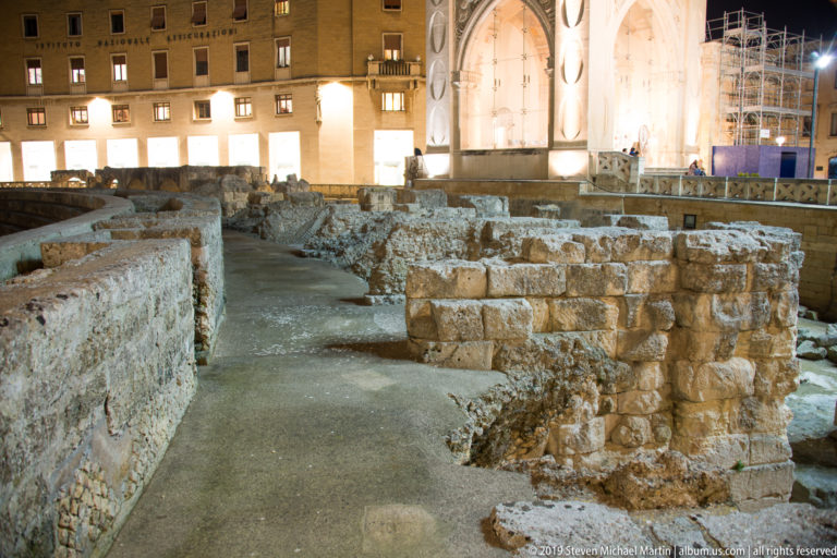Low excavated stone walls and passages illuminated at night within the Roman amphitheatre archaeological area in central Lecce.