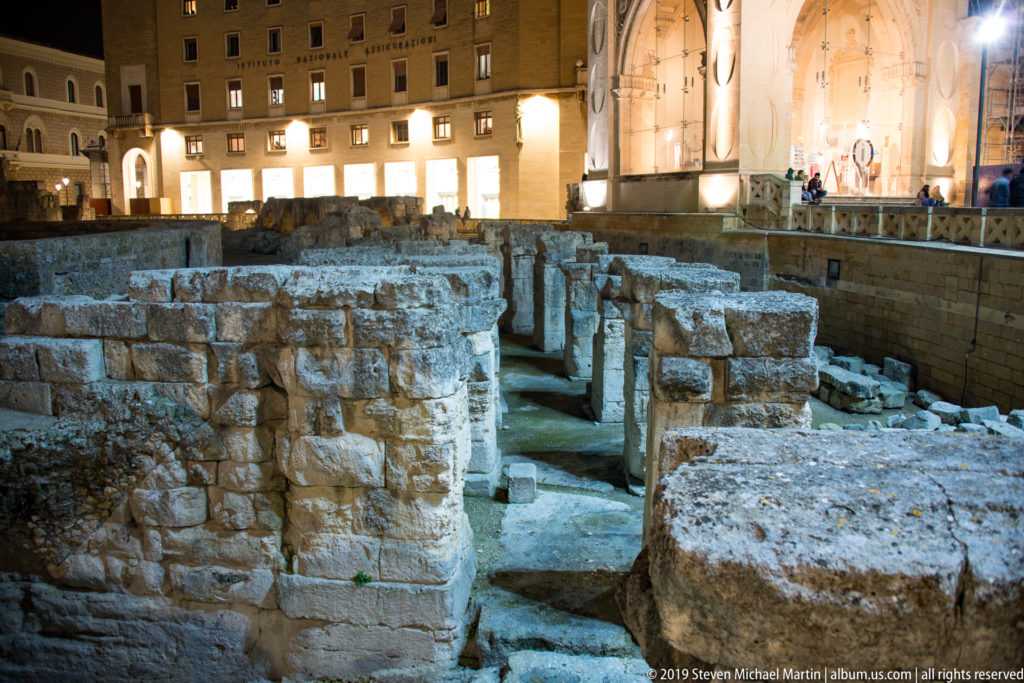 Excavated walls and stone blocks of Lecce’s Roman amphitheatre with illuminated buildings of Piazza Sant’Oronzo beyond.