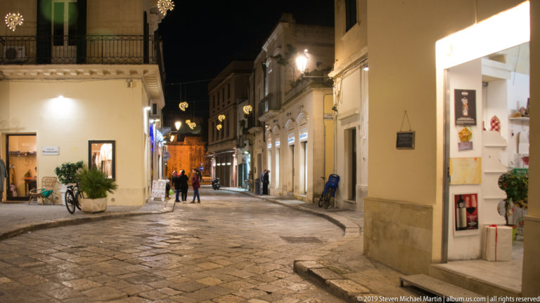Curving stone-paved street lined with illuminated shops and historic façades, with pedestrians, bicycles, and hanging lights at night.