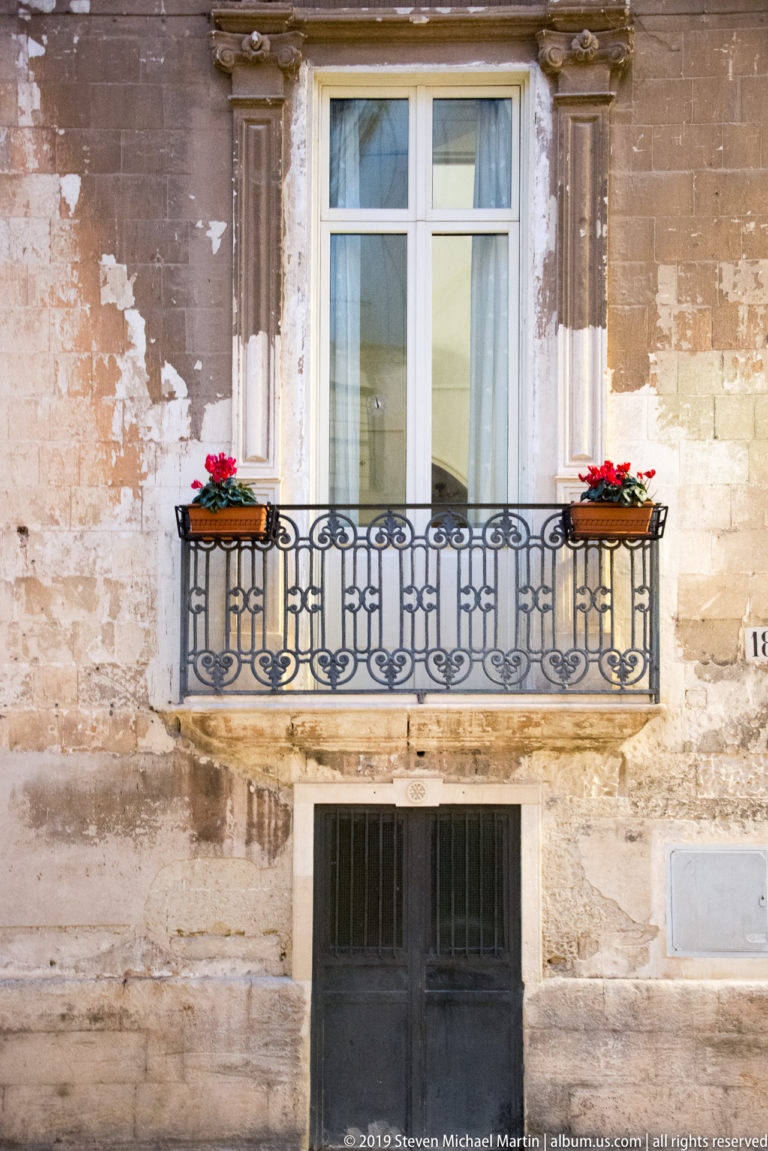 Black wrought-iron balcony with red flower boxes beneath a white window on a weathered pale-stone façade.