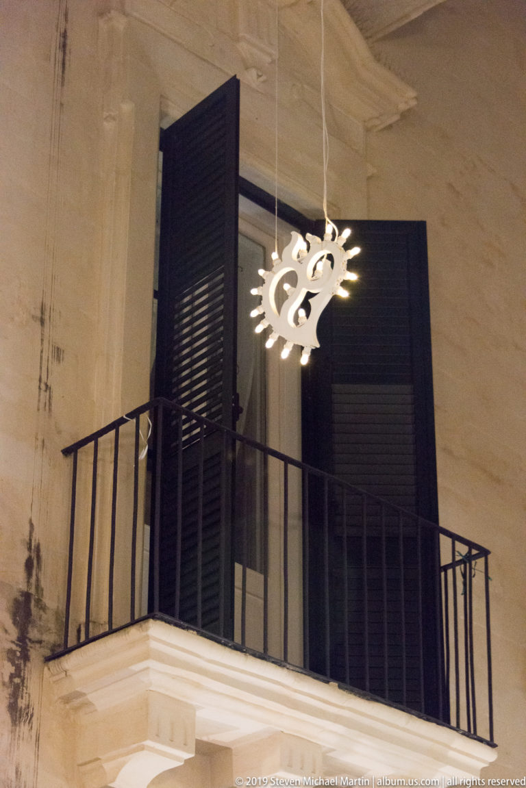 Glowing hanging ornament beside a small iron balcony with open dark shutters on a pale stone façade.