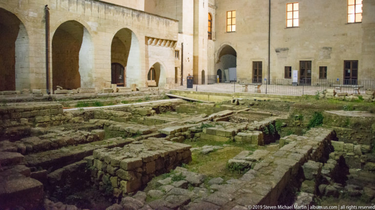 Excavated stone foundations and wall lines filling a courtyard bordered by pale masonry walls, arches, and lit windows.
