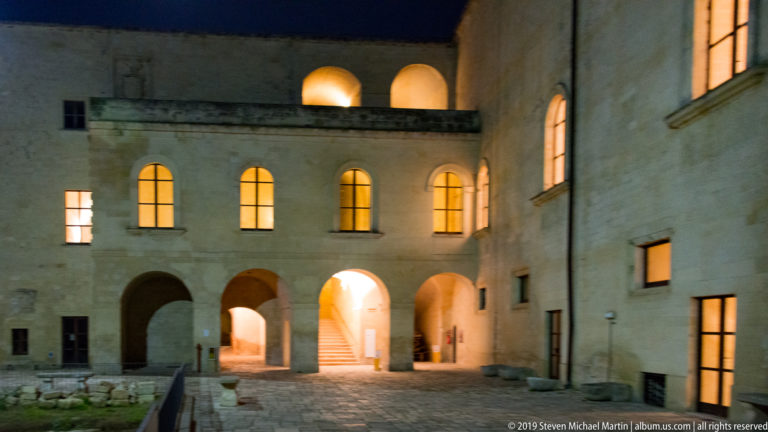 Stone courtyard at night with glowing arched openings, windows, stairs, and passages arranged across multiple levels.