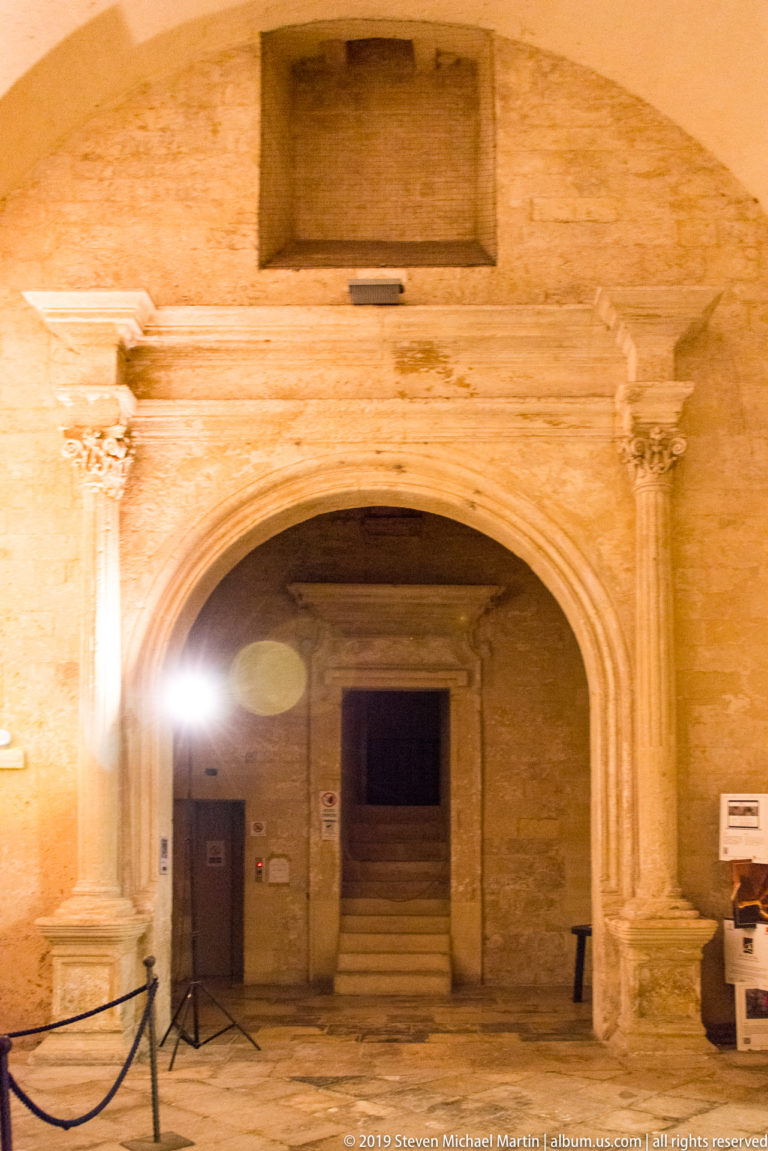 Large pointed stone arch with slender side columns opening to a recessed doorway and steps in a historic masonry interior.