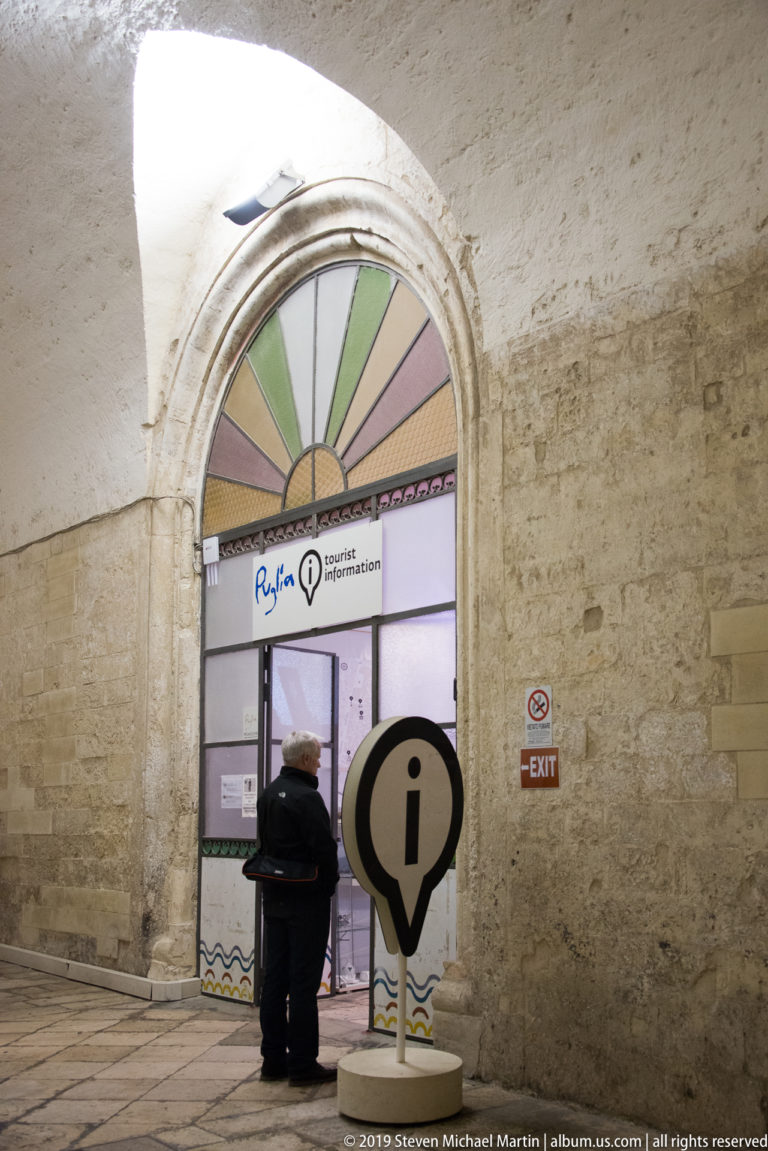 Visitor standing at a tourist-information entrance beneath a stone vault, with a large information symbol in the foreground.