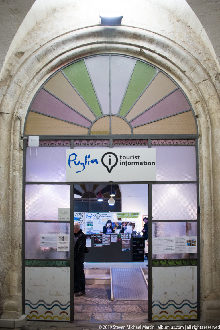 Tourist-information office set within a large stone arch, with colored fanlight panes, illuminated signage, screens, and a visitor inside.