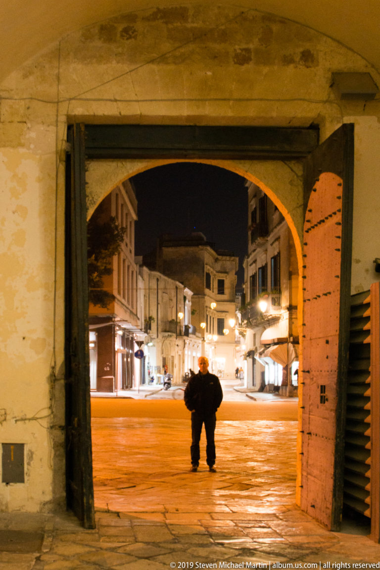 Silhouetted person beneath a dark arched passage facing a brightly lit stone-paved street at night.