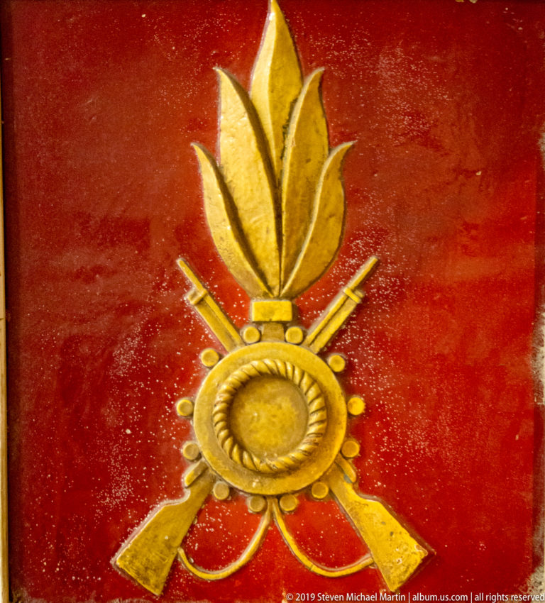 Gold-colored flaming grenade emblem with circular center and crossed weapon forms mounted against a red background.