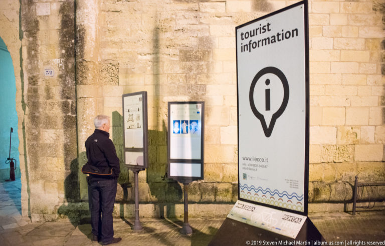 Person reading illuminated information panels beside a large freestanding tourist-information sign against a historic stone wall.