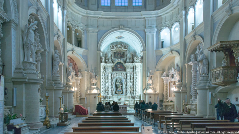Bright Baroque church nave with sculpted piers, side altars, visitors, high windows, and an elaborate white-stone high altar.