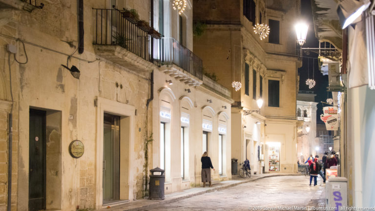Narrow stone-paved shopping street at night with pedestrians, illuminated storefronts, balconies, bicycles, and hanging decorative lights.