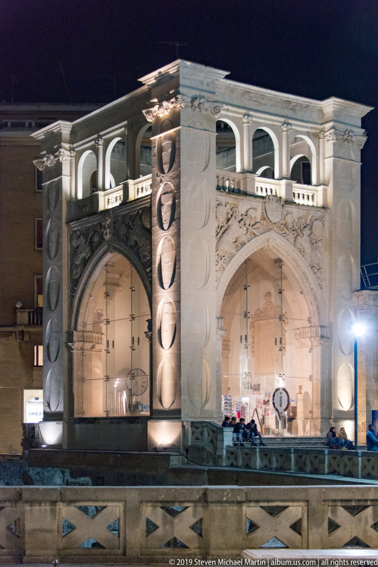Bright modern pavilion with two large pointed arches, open upper gallery, and circular relief motifs illuminated at night.