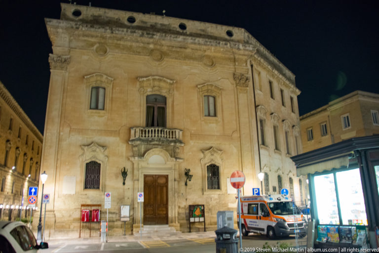 Illuminated historic corner building with central balcony and arched doorway, with an ambulance parked along the street.