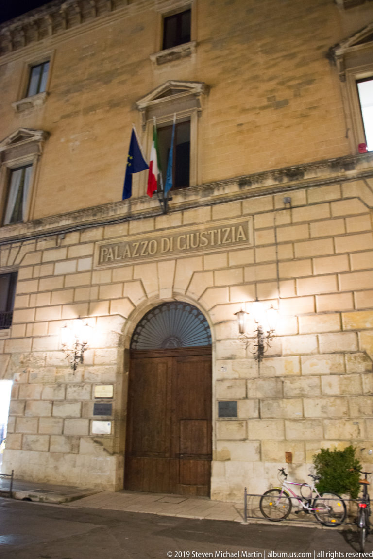 Palazzo di Giustizia in Lecce at night with arched wooden entrance, identifying inscription, Italian and European flags, and bicycles.