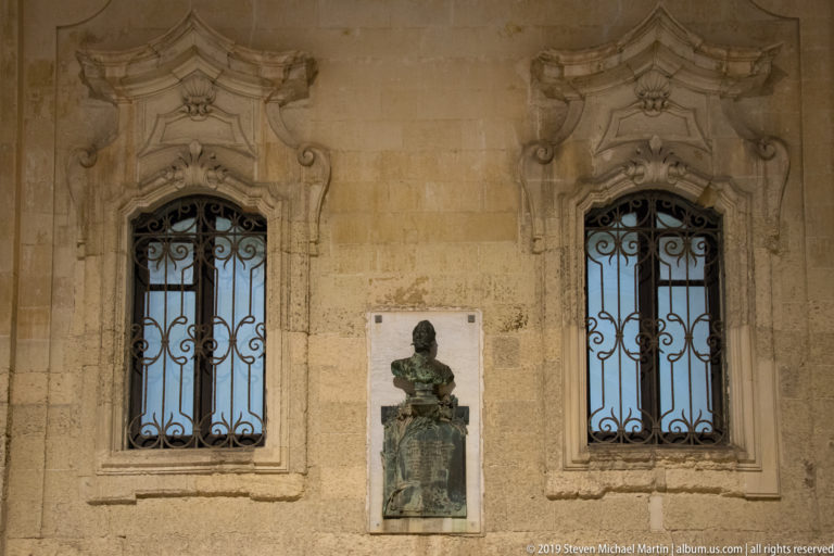 Bronze bust on a commemorative plaque centered between two iron-grilled windows with ornate carved stone surrounds.