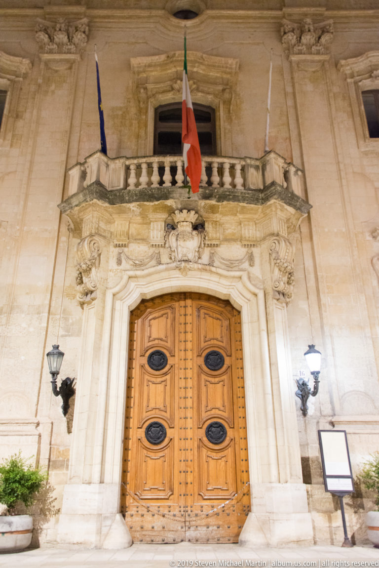 Formal stone entrance with carved wooden double doors, ornate surround, balcony, wall lamps, and an Italian flag overhead.