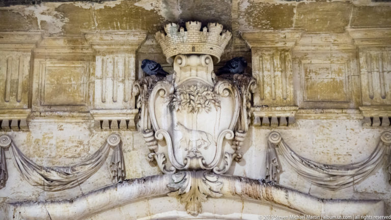 Crowned carved-stone cartouche surrounded by scrollwork, draped swags, moldings, and weathered architectural ornament.