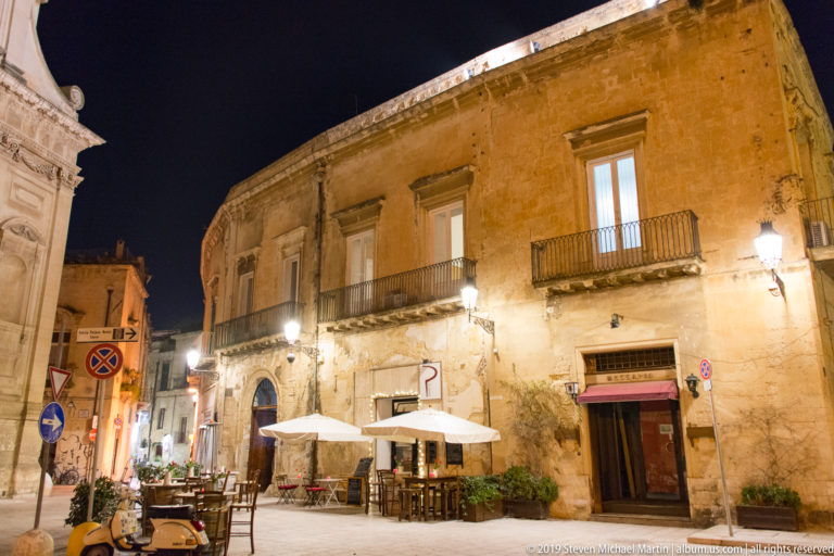 Long illuminated stone palazzo façade with balconies and tall windows above café tables, umbrellas, scooters, and street signs.