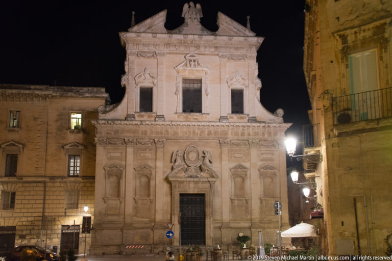 Illuminated Baroque church façade with ornate portal and upper windows, framed by neighboring stone buildings at night.