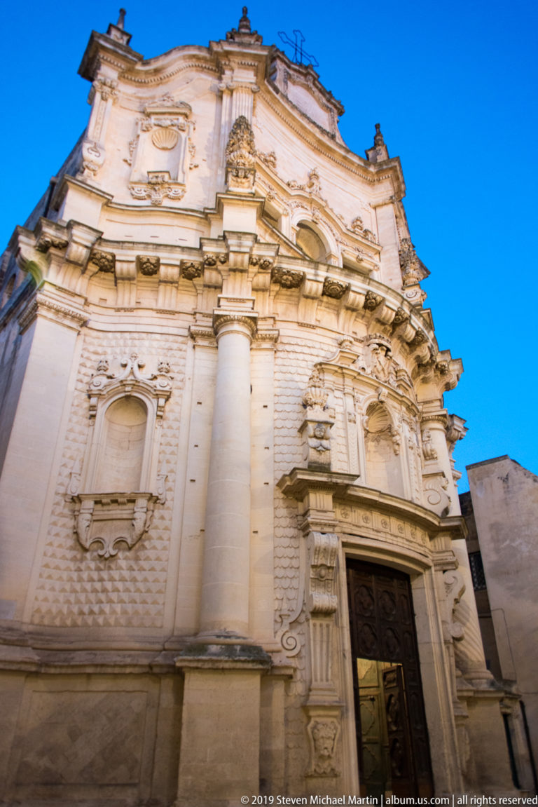 Oblique upward view of an illuminated Baroque church façade with giant columns, niches, carved doorway, and curving upper profile.