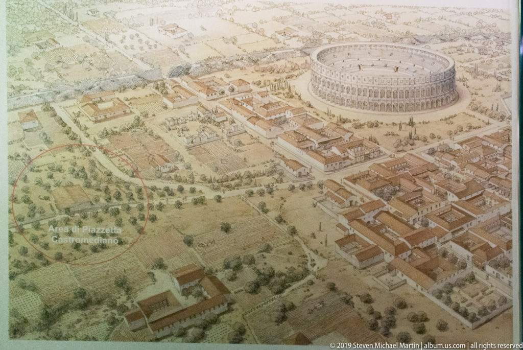 Illustrated aerial reconstruction of ancient Lecce with a large oval Roman amphitheatre surrounded by streets, buildings, and open land.