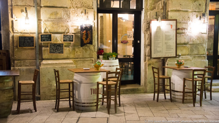 Outdoor café tables and chairs beside a warmly illuminated stone façade with plaques, menu boards, and a glazed doorway.