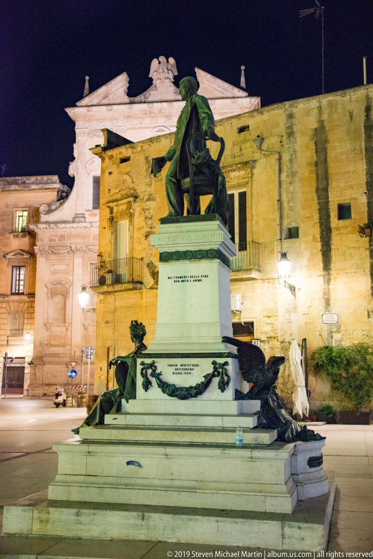 Bronze seated figure atop a tall stone pedestal with decorative metalwork, set before illuminated historic buildings at night.