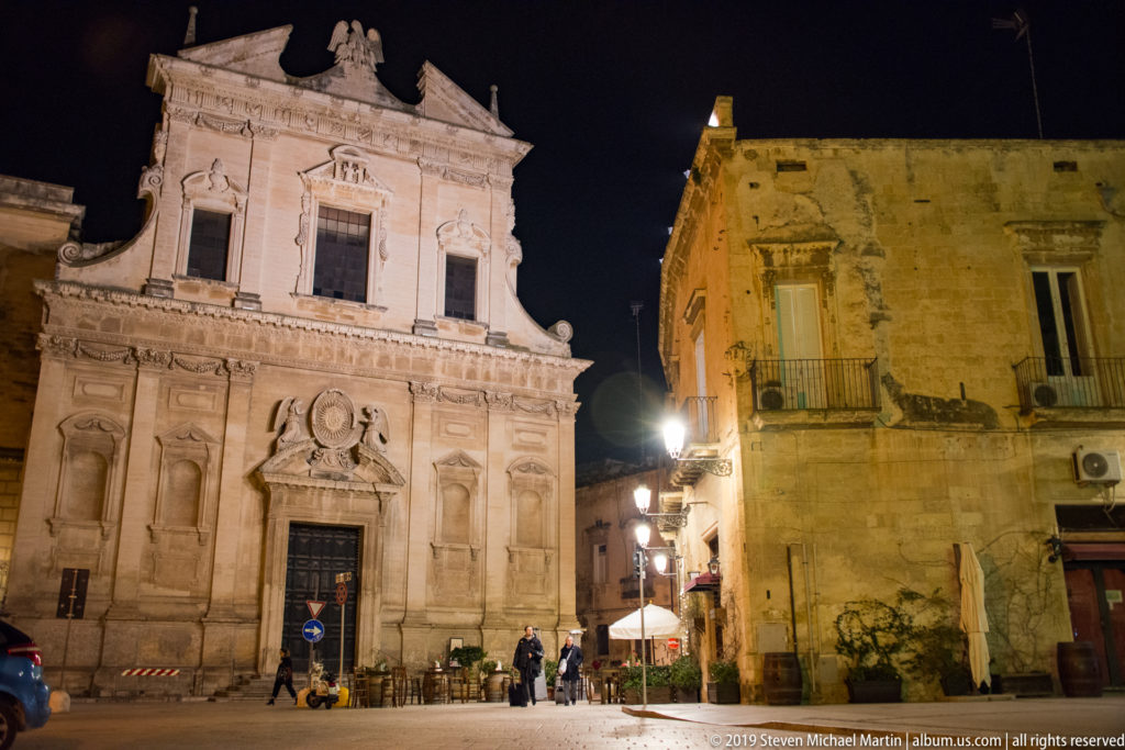 Oblique night view of ornate church beside a plain stone building, with pedestrians and café umbrellas near the entrance.