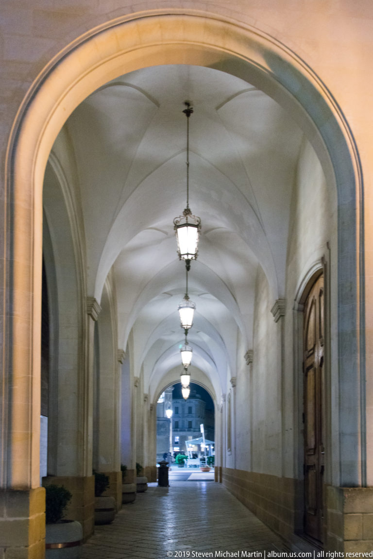 Long vaulted passage with evenly spaced hanging lanterns, wooden doors, stone paving, and a bright street opening ahead.