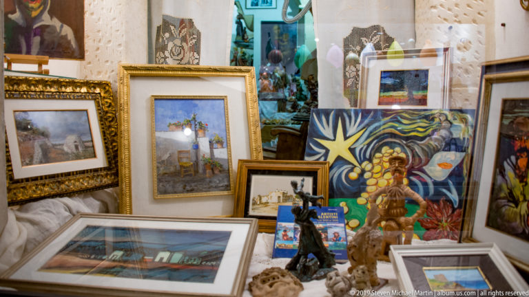 Storefront filled with framed paintings, colorful artwork, small sculptures, ceramics, and reflections in the display glass.