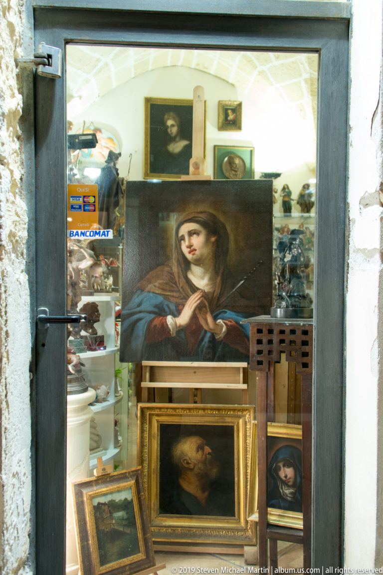Shop window crowded with framed religious paintings, including a large portrait of a veiled woman and smaller devotional works.