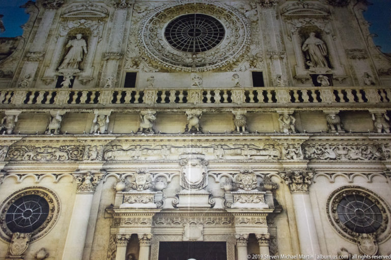 Upward view of ornate church façade with large rose window, statues in niches, balustrade, and dense limestone carving.