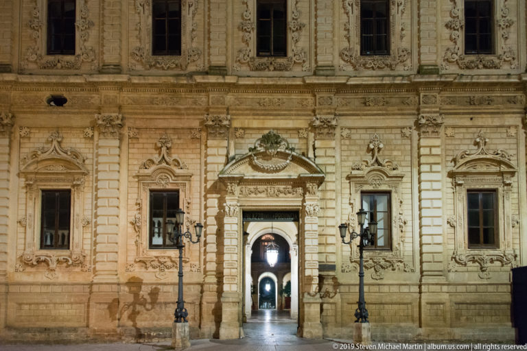 Illuminated Palazzo della Provincia façade with ornate central portal, tall windows, paired lamps, and passage visible beyond.